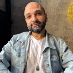 Mavi cins şalvar və açıq mavi köynək geyinmiş bir kişi, arxa planda açıq tonlu divar ilə oturmuş.